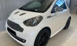 Biały Aston Martin Cygnet z czarnymi felgami i chromowanymi detalami, prezentujący się elegancko w garażu.