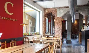 Interno di un locale con tavoli in legno e mattoni a vista. Sulla parete rossa, il logo "C CARROBIOLO" e una vetrina con tante bottiglie di birra.