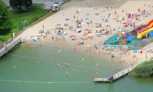 Wakacyjny dzień nad jeziorem margonińskim. Ludzie opalają się na plaży, kąpią w wodzie i korzystają ze zjeżdżalni wodnych.