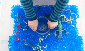 Kinderhände erkunden eine Kiste voller blauer Wasserperlen mit Spielzeug-Echsen und Schlangen. Tolle Wasserperlen Ideen für sensorisches Spiel!