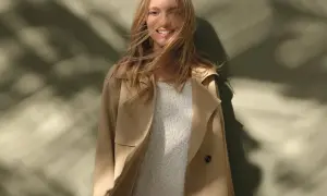 Uśmiechnięta blondynka w beżowym trenczu i swetrze, z rozwianymi włosami. Inspiracja, jak nosić trencz w wiosenny dzień.