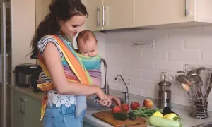 Mamma förbereder mat vid amning i köket, med en bebis i bärsjal. Grönsaker ligger på bänken.