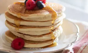 Pancakes senza uova dorati, impilati e inzuppati di sciroppo, guarniti con lamponi e mirtilli freschi.