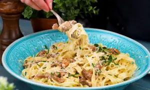 En gaffel lyfter en portion krämig pasta med kantareller, toppad med parmesan och färska örter i en vacker blå skål.