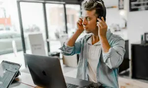 A young man, likely one of the new generation of music managers, adjusts his headphones while working on his laptop in a cafe.