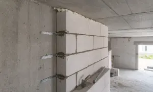 Pokazano, jak łączyć ściany działowe z nośnymi za pomocą metalowych kątowników i pianki montażowej. Narzędzia budowlane leżą na murze.