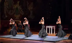 Cuatro bailarinas de flamenco, con vestidos negros voluminosos, ejecutan pasos apasionados. Un hombre aplaude al fondo.