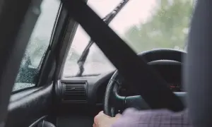 Widok z wnętrza samochodu podczas jazdy w deszczu. Krople na szybie, wycieraczki pracują. Kierowca trzyma kierownicę.