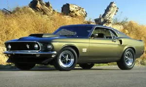 Ein goldener amerikanischer Oldtimer, ein Ford Mustang Boss 429, steht vor einer trockenen, grasbewachsenen Böschung mit Baumstämmen.