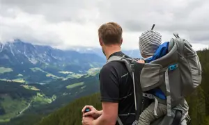 Mann mit Kind auf dem Rücken in einer Kraxe, blickt auf bergige Landschaft.