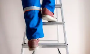 Osoba w niebieskich spodniach roboczych wspina się po drabinie, przestrzegając **drabiny wymagania bhp**.