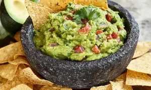 En skål med krämig guacamole, perfekt för ditt nästa guacamole recept. Serveras med tortillachips och en halv avokado i bakgrunden.