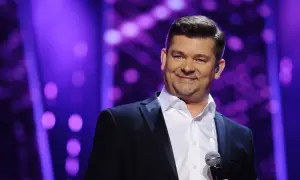 Uśmiechnięta gwiazda w czarnym garniturze, z mikrofonem w dłoni, na fioletowym tle. Jego akcent jest wyraźny, a tekst piosenki płynie.
