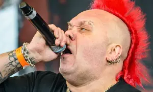 Wokalista z irokezem śpiewa do mikrofonu. Widać tatuaże, kolczyki i opaskę festiwalową. Jego styl sugeruje, że nie dał się łatwo wykorzystać.