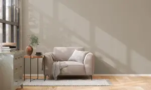 Un sillón beige con manta y cojín, junto a una mesita auxiliar con jarrón y taza. La pared tiene unos colores de pintura neutros, con sombras de luz.