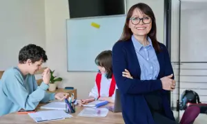 Nauczycielka z uśmiechem nadzoruje zajęcia z wychowawcą. Uczniowie pracują przy biurku, a na tablicy widać rozkład materiału.