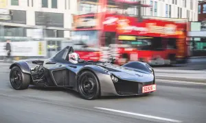 Niesamowity BAC Mono pędzi ulicą, mijając rozmazany czerwony autobus i budynki.