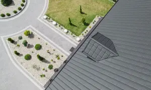 Nowoczesny dach pulpitowy z szarej blachy, z widocznym oknem dachowym. Obok zadbany ogród z brukowanymi ścieżkami i niską roślinnością.