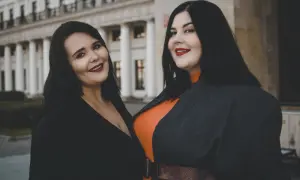 Dwie uśmiechnięte kobiety, uosobienie body positivity, pozują przed budynkiem z kolumnami.