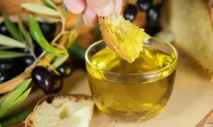 Un pezzo di pane intinto in olio dorato, con olive e rami d'ulivo sullo sfondo, evoca le antiche origini olio di oliva.
