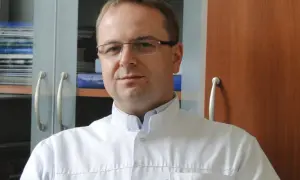Portret mężczyzny w okularach i białym fartuchu lekarskim, na tle regałów z książkami.