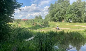 Osoba na motocyklu crossowym jedzie po szutrowej drodze wśród zieleni, obok stawu i drewnianego mostku.