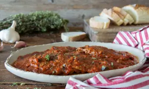 Herzhaftes Carne alla Pizzaiola in Tomatensauce, garniert mit Kräutern, neben Knoblauch, Brot und einem rot-weiß gestreiften Tuch.