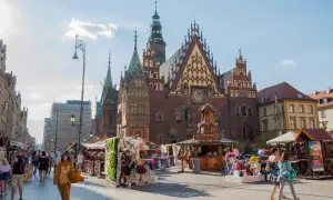 Tłum ludzi spaceruje po Rynku we Wrocławiu, gdzie można pojechać w niedzielę. W tle widać piękną architekturę i kramy z kwiatami.