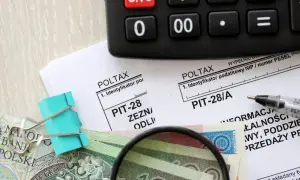 Kalkulator, formularze PIT, pieniądze i lupa na drewnianym tle.