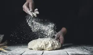 Mjöl dammas över deg. En hand strör mjöl över en deg på ett träbord, med veteax i bakgrunden. Råg vs vete – valet av mjöl påverkar bakningen.