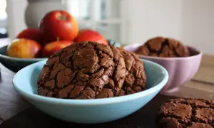 Mörka, sega kladdkake cookies i en ljusblå skål, med äpplen i bakgrunden. En annan skål med cookies syns i bakgrunden.