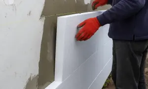 Pracownik w czerwonych rękawicach montuje płyty styropianowe do ściany, rozpoczynając ocieplanie elewacji.
