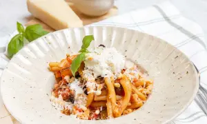 En tallrik med pasta alla norma, toppad med ricotta och basilika. Bredvid syns en bit parmesanost och en skål med pinjenötter.