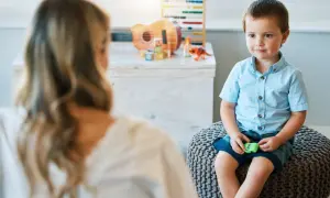 Chłopiec w niebieskiej koszuli siedzi na pufie, rozmawiając z psychologiem. W tle zabawki.