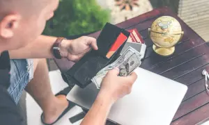 Mężczyzna liczy pieniądze w portfelu, przygotowując się do podróży. Na stole leży globus, paszport i laptop, sugerując potrzebę finansowego assistance.