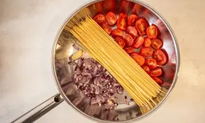 Zutaten für ein One-Pot-Gericht: Spaghetti, gehackte rote Zwiebeln, Kirschtomaten und eine Knoblauchzehe in einer Pfanne.