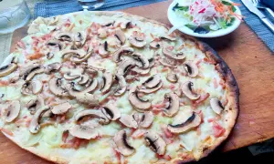 Ein köstlicher Straßburger Flammkuchen mit Pilzen und Speck, serviert auf einem Holzbrett, daneben ein kleiner Salat.