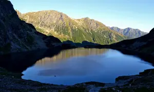Górskie **rysy z Morskiego Oka** odbijają się w spokojnej tafli wody, otoczonej skalistymi zboczami Tatr.