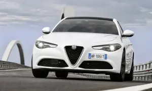 Biała Alfa Romeo Giulia Quadrifoglio z czarnym dachem pędzi po drodze z mostami w tle.