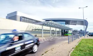 Taksówka pędzi przed nowoczesnym terminalem lotniska Poznań Ławica. W tle zielony autobus i inne pojazdy.