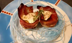 Zwei Eier mit Speck, kunstvoll in einem Nest aus blauen Fäden auf einem Teller angerichtet.