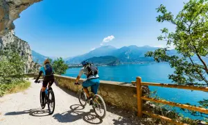 Ciclisti su una strada panoramica lungo il Lago di Garda, con montagne sullo sfondo.