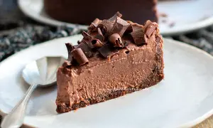 Una fetta di goloso cheesecake al cioccolato, guarnita con riccioli di cioccolato fondente, su un piatto bianco con un cucchiaino.