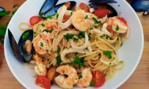 Talerz spaghetti z owocami morza: krewetkami, małżami i kalmarami, ozdobiony pomidorkami i natką pietruszki.