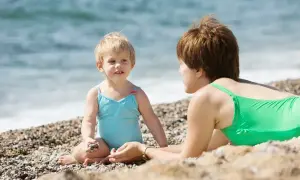 Mama i dziecko cieszą się wspólnym czasem nad morzem z dziećmi. Dziewczynka w turkusowym kostiumie bawi się kamykami, a mama w zielonym kostiumie leży obok.