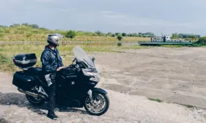 Motocyklista w kasku i kurtce stoi obok czarnego motocykla nad kanałem z łodzią w tle.
