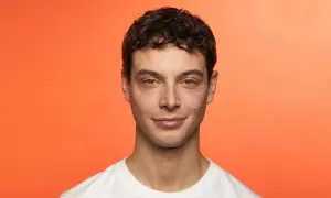 Hombre joven con cabello castaño rizado, luciendo una permanente ondulada. Sonríe levemente ante un fondo naranja vibrante.