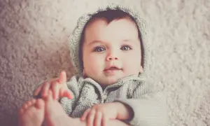 Bebé con ojos grandes y azules, vistiendo un gorro y suéter tejidos. Podría llamarse Gabriel, un nombre con g.