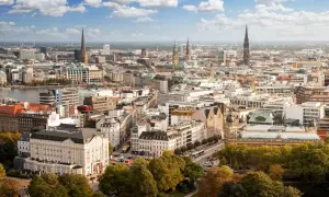 Blick von einer **Aussichtsplattform Hamburg** über die Stadt mit ihren Kirchtürmen, dem Hafen und grünen Bäumen im Vordergrund.