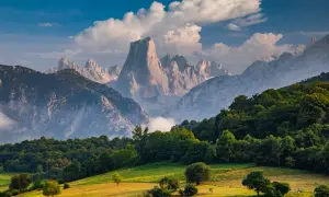 Majestuosas montañas de los Picos de Europa se alzan tras un prado verde y un bosque frondoso, bajo un cielo azul con nubes algodonosas.